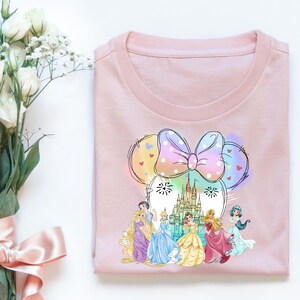 May include: Pink t-shirt with a colorful graphic of a Mickey Mouse head with a bow and a castle in the background. The castle is surrounded by six Disney princesses.