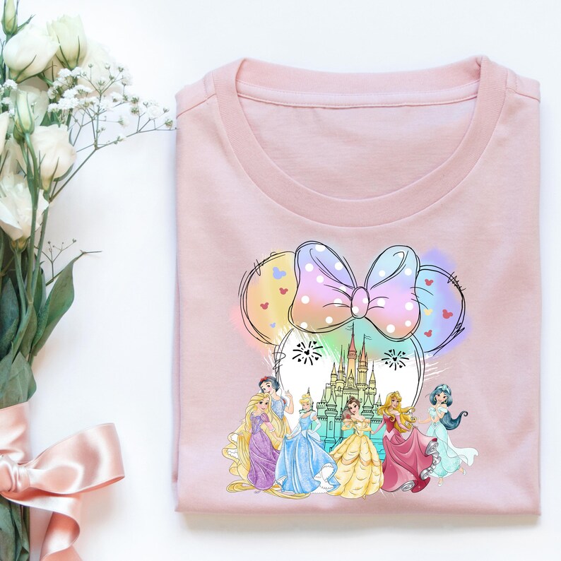 May include: Pink t-shirt with a colorful graphic of a Mickey Mouse head with a bow and a castle in the background. The castle is surrounded by six Disney princesses.