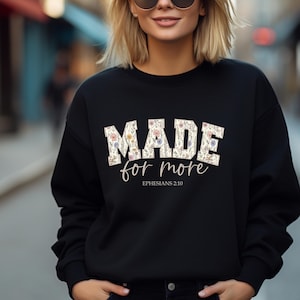 May include: Black crew neck sweatshirt featuring the text "MADE for more" in a floral font. The words "Ephesians 2:10" are printed below. The sweatshirt is a casual style.