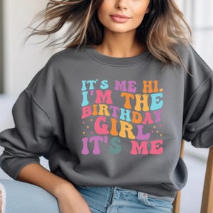 May include: A gray sweatshirt with the text "It's me hi. I'm the birthday girl. It's me" in colorful, retro-style lettering.