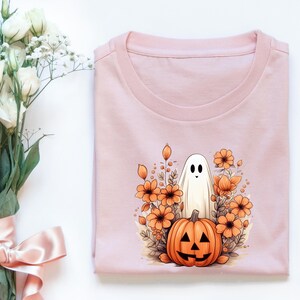 May include: A pink t-shirt with a white ghost and an orange pumpkin with a jack-o'-lantern face in a field of orange flowers.