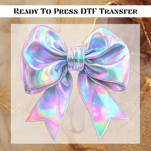 May include: A holographic iridescent bow with a pink, blue, and green colour scheme. The bow is ready to be pressed onto a fabric surface using a heat press.