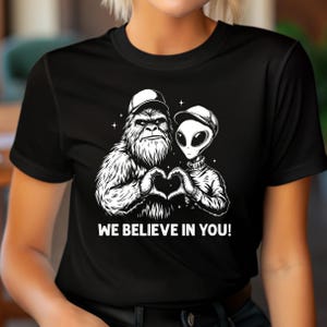 May include: Black t-shirt with a white graphic of a Bigfoot and an alien making a heart shape. The text "WE BELIEVE IN YOU!" is printed below. The design is in black and white, featuring a whimsical illustration.