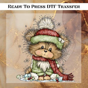 May include: A DTF transfer featuring a cartoon bear wearing a red and green winter hat and scarf. The bear is dressed in a green coat and is standing in snow. The text "READY TO PRESS DTF TRANSFER" is at the top.