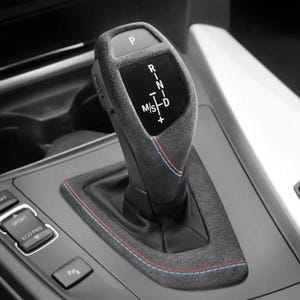 May include: A close-up of a black leather car gear shifter with a silver trim. The shifter has a black leather boot with red and blue stitching. The shifter has the letters "P", "R", "N", "M/S", "D", and "+" printed on it.