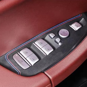 May include: Close-up of a car door panel with a burgundy leather interior. The panel features silver-colored window and lock controls, a central control dial, and a black trim with blue and red stitching.