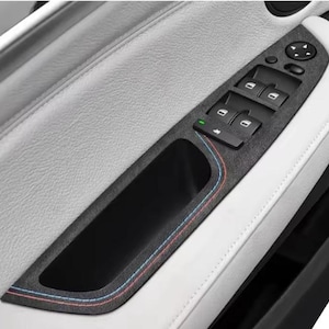 May include: A car door interior detail showing a black control panel with window switches and a door handle. The panel is set against a white leather interior with a textured surface. Red and blue stitching accents the black panel.