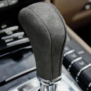 Puede incluir: Primer plano de una palanca de cambios de coche de color gris, con aspecto de ante. El pomo tiene un diseño redondeado y ergonómico con una base plateada. El fondo muestra detalles del interior del coche.