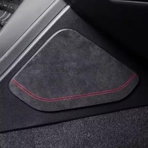 Könnte beinhalten: Ein dunkelgraues, Wildleder-ähnliches Innenraumteil für ein Auto mit roter Naht. Das Panel ist in einen schwarzen Kunststoffrahmen eingesetzt, mit einem Blick auf andere Autoteile im Hintergrund.