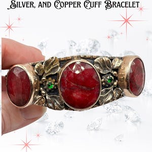 Op de afbeelding: Vintage ambachtelijke manchetarmband met grote robijnstenen, smaragdaccenten en goudkleurig metaalwerk. De armband heeft een bloemenontwerp en is gemaakt van zilver en koper. De tekst op de afbeelding luidt: "VINTAGE ARTISAN RUBY, EMERALD, SILVER, AND COPPER CUFF BRACELET."