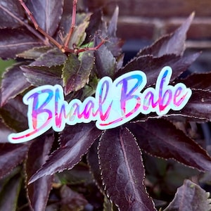May include: A sticker featuring the text Bhaal Babe in a stylized, cursive font. The lettering displays a vibrant, multi-colored holographic gradient with shades of pink, blue, and purple, outlined in a thin white border against a dark background.