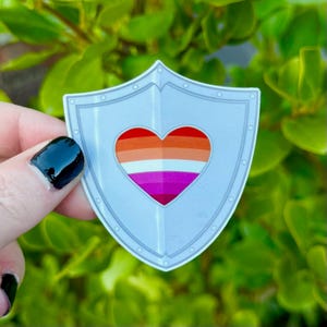 May include: A shield-shaped sticker featuring a central heart graphic filled with horizontal stripes in shades of orange, white, and magenta. The shield has a light gray metallic appearance with a defined border and small circular rivet details.