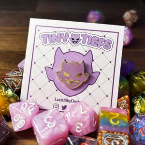 May include: A gold-colored enamel pin featuring a purple cartoon tiefling head with horns and a mischievous grin. The pin is on a white card with the text "Tiny Tiefs" and "LucidSkyDesign" in purple and black lettering.