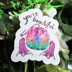 May include: A holographic sticker with a cartoon creature design. The creature has a brain-shaped head in pink, blue, and green, and purple feet. The text "you're beautiful" is written in black script. The sticker has a sparkly effect.