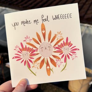 May include: A square greeting card with a floral design. The card features a central flower with a face, surrounded by other flowers in shades of orange and pink. The text "you make me feel WHEEEEE" is written at the top.