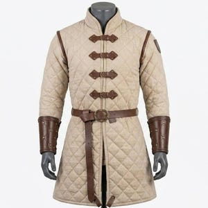 Könnte beinhalten: Beige gesteppter mittelalterlicher Gambeson mit braunen Lederakzenten. Mit hohem Kragen, dekorativen Lederverschlüssen und passendem Gürtel. Die Ärmel haben braune Lederbündchen. Ein historisches Kleidungsstück.