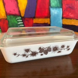 May include: A rectangular white casserole dish with a clear glass lid. The dish features a brown pinecone and branch design. The dish is approximately 10 inches long, 6 inches wide, and 3 inches deep.