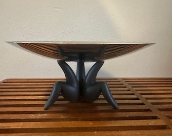 May include: A silver serving bowl with a wide, shallow, round shape sits on a black, stylized, human-shaped stand with legs. The stand is on a wooden surface.