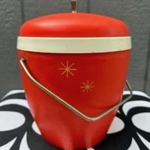 May include: A vintage red ice bucket with a white band and gold starburst designs. It has a matching red lid with a small gold handle and a gold metal handle for carrying. The bucket is approximately 10 inches tall.