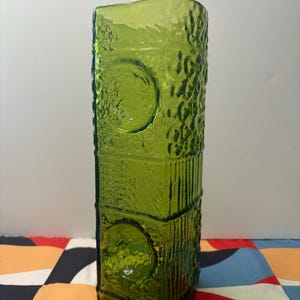 May include: A tall, green glass vase with a textured surface and two circular cutouts. The vase has a rectangular shape and is made of thick glass.