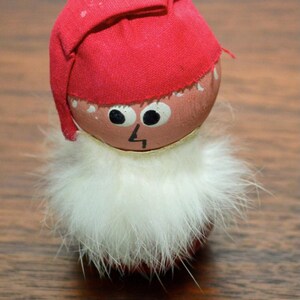 May include: A small, red and white Christmas gnome figurine with a white fur beard and a red hat. The gnome has a painted face with black eyes and a small, black nose.
