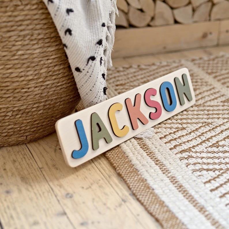 K&ouml;nnte beinhalten: Ein Holzpuzzle mit dem Namen "JACKSON" in bunten Buchstaben. Die Buchstaben sind blau, gr&uuml;n, gelb, rosa und blau. Das Puzzle liegt auf einer hellen Holzoberfl&auml;che, mit einem gewebten Teppich und einem Korb im Hintergrund.