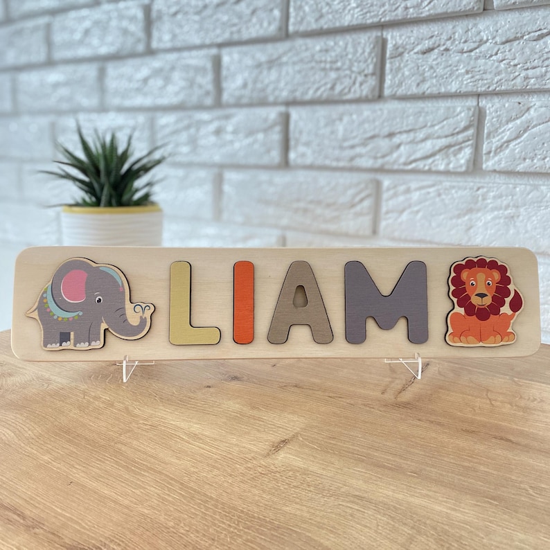 K&ouml;nnte beinhalten: Holz-Namenspuzzle mit dem Namen "LIAM" in bunten Buchstaben. Mit einer Elefanten- und L&ouml;wen-Illustration. Das Puzzle ist aus hellem Holz gefertigt und f&uuml;r Kinder konzipiert.