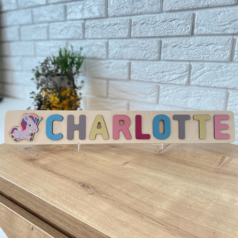 K&ouml;nnte beinhalten: Holzpuzzle mit dem Namen "CHARLOTTE" in bunten Buchstaben. Auf der linken Seite befindet sich ein Cartoon-Einhorn. Das Puzzle liegt auf einer Holzoberfl&auml;che mit einer wei&szlig;en Backsteinmauer im Hintergrund.