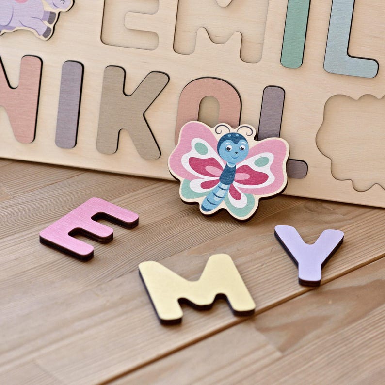 K&ouml;nnte beinhalten: Holz-Namenspuzzle mit bunten Buchstaben und einem schmetterlingsf&ouml;rmigen Teil. Die Buchstaben sind in verschiedenen Pastellfarben gehalten, mit einem rosa "E", einem gelben "M" und einem lila "Y". Der Schmetterling hat rosa, blaue und gr&uuml;ne Akzente.