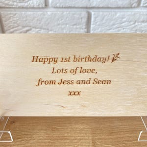 K&ouml;nnte beinhalten: Helle Holzplakette mit graviertem Text. Der Text lautet: "Happy 1st birthday! Lots of love, from Jess and Sean xxx". Die Plakette wird von transparenten Acrylst&auml;ndern getragen, und der Hintergrund ist eine wei&szlig;e Backsteinmauer.