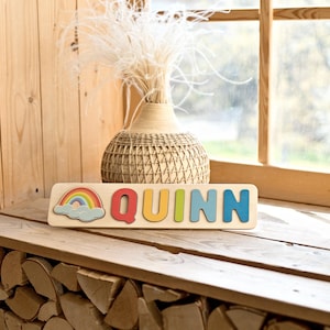 K&ouml;nnte beinhalten: Ein Holzpuzzle mit dem Namen "QUINN" in bunten Buchstaben. Links befindet sich eine Regenbogen- und Wolkengrafik. Das Puzzle liegt auf einer Holzoberfl&auml;che, im Hintergrund eine dekorative Vase und ein Fenster.