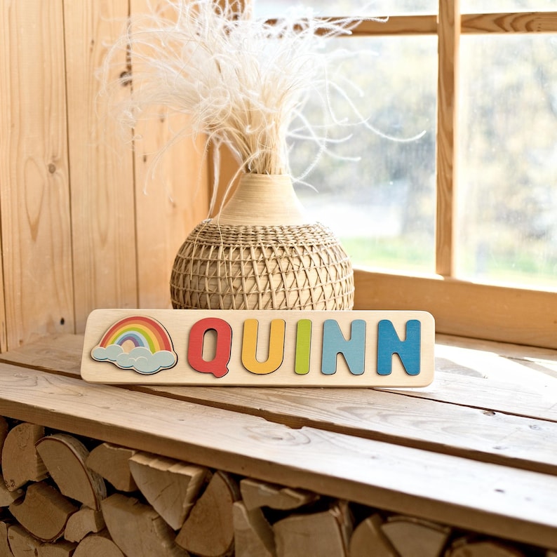 K&ouml;nnte beinhalten: Ein Holzpuzzle mit dem Namen "QUINN" in bunten Buchstaben. Links befindet sich eine Regenbogen- und Wolkengrafik. Das Puzzle liegt auf einer Holzoberfl&auml;che, im Hintergrund eine dekorative Vase und ein Fenster.