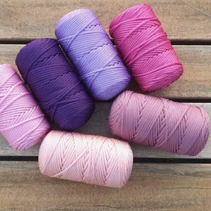 Könnte beinhalten: Draufsicht auf sieben Spulen farbenfrohen Makramee-Garns in Rosa-, Lila- und Lavendeltönen. Die Spulen sind auf einer Holzoberfläche angeordnet und zeigen die Vielfalt der Farben und Texturen.