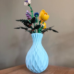 Könnte beinhalten: Eine hellblaue Keramikvase mit einem strukturierten Diamantenmuster hält einen Strauß bunter Lego-Blumen. Die Blumen umfassen eine weiße und violette Blume, eine gelbe Blume und eine blaue Blume.