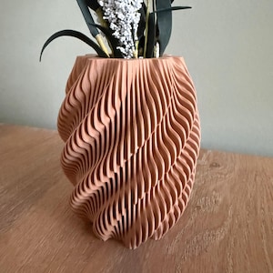 Op de afbeelding: Een perzikkleurige, ingewikkeld gesneden houten vaas met een wervelend, gelaagd ontwerp. De vaas bevat witte en groene kunstbloemen. De vaas staat op een houten oppervlak.