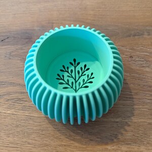 May include: A round, turquoise-colored planter with a ribbed exterior and a black tree design on the interior base. The planter is made of a solid material and sits on a wooden surface.
