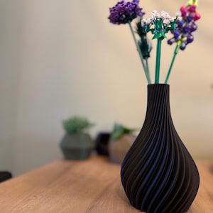 Könnte beinhalten: Eine schwarze, gewellte Vase mit strukturierter Oberfläche hält einen Strauß aus lila, weißen und rosa Blumen. Die Vase steht auf einem Holztisch.