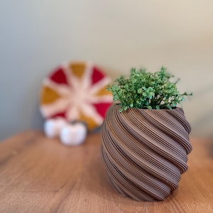 Op de afbeelding: Een bruine, spiraalvormige plantenpot met een klein groen plantje erin. De pot staat op een houten tafel.