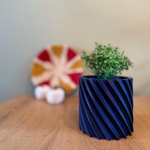 Op de afbeelding: Een donkerblauwe plantenpot met een spiraalvormig ontwerp bevat een kleine groene plant. De pot staat op een houten tafel.