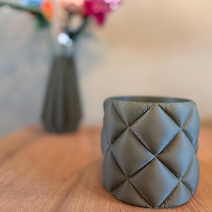 May include: A grey, quilted-patterned plant pot is on a wooden surface. The pot has a cylindrical shape and a diamond pattern. In the background, a vase with colourful flowers is slightly out of focus.