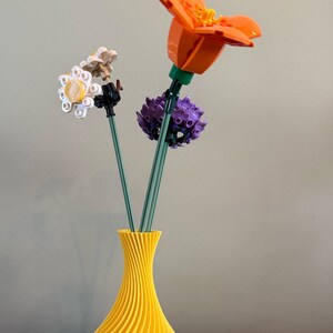 Könnte beinhalten: Eine gelbe Keramikvase mit einem wellenförmigen Design hält drei bunte Lego-Blumen. Die Blumen sind orange, weiß und lila.
