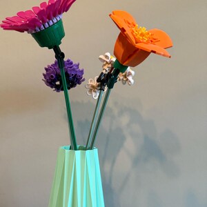 Op de afbeelding: Een lichtgroene origami-vaas met drie kleurrijke Lego-bloemen. De bloemen zijn roze, oranje en paars.