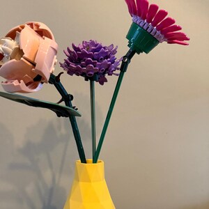 Könnte beinhalten: Eine gelbe geometrische Vase mit drei Blumen aus Lego-Steinen. Die Blumen sind rosa, lila und grün.