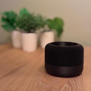 May include: A black cylindrical smart speaker with a mesh grille on the top and bottom. The speaker is sitting on a wooden table.