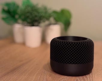 May include: A black cylindrical smart speaker with a mesh grille on the top and bottom. The speaker is sitting on a wooden table.