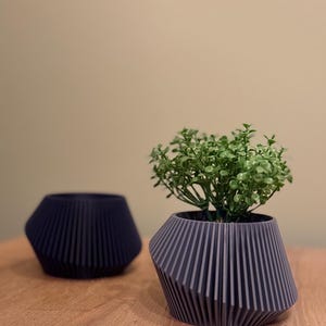 May include: Two grey geometric planters, one with a small green plant. The planters have a ribbed design and are on a wooden surface.