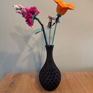 May include: A black vase with a geometric pattern holds three artificial flowers. The flowers are pink, orange, and white.