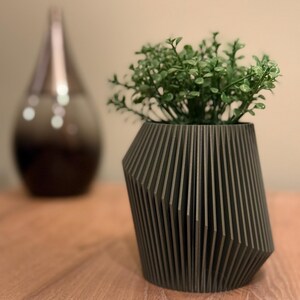 May include: A dark grey geometric planter with a green succulent plant inside. The planter has a unique design with vertical lines and a slightly angled shape.