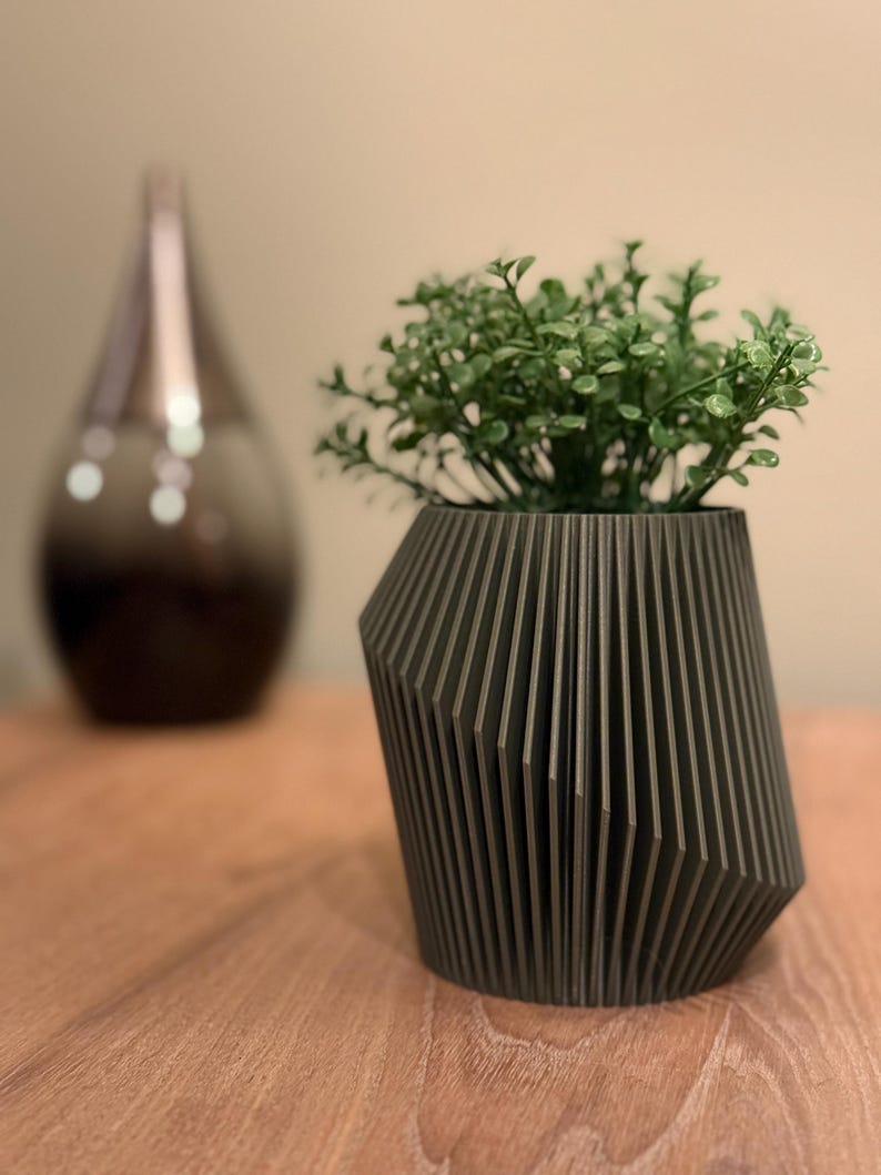May include: A dark grey geometric planter with a green succulent plant inside. The planter has a unique design with vertical lines and a slightly angled shape.