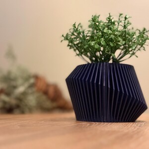 May include: A dark blue, geometric planter with a ribbed design, holding a small green plant.